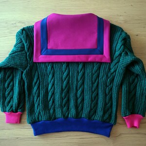 Peut inclure: Un pull vert en tricot torsadé avec un grand col magenta bordé de bleu. Les poignets sont roses et l'ourlet inférieur est bleu. Le pull est présenté sur une surface en bois brun clair.