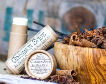 May include: Two brown cardboard tubes of cinnamon spice lip balm, one with a label that reads "Cinnamon Spice All Natural Lip Balm 0.45 fl oz" and the other with a label that reads "Cinnamon Spice Lip Balm 1.0 fl oz". A wooden bowl filled with cinnamon sticks and star anise sits on a burlap surface.