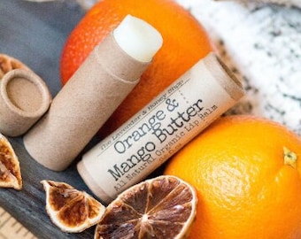 May include: A brown cardboard tube lip balm with a white label that reads "The Lavender & Honey Shop Orange & Mango Butter All Natural Organic Lip Balm". The lip balm is sitting on a wooden surface with dried orange slices and a whole orange.