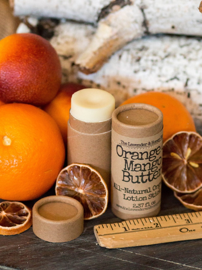 May include: A brown cardboard tube with a white label that reads "The Lavender & Honey Shop Orange Mango Butter All-Natural Organic Lotion Stick 2.37 fl oz www.thelavenderandhoneyshop.com". The tube is sitting on a wooden surface with oranges, dried orange slices, and a wooden ruler.