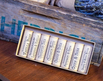 May include: A set of eight small, cylindrical soap bars in a white box. The soaps are labeled with different scents, including Vanilla Bean, Lavender & Vanilla, Unscented Shea, Cinnamon Spice, Orange & Cocoa, Peppermint & Cocoa, Cocoa & Shea, and Orange & Mango Butter.