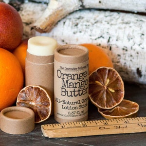 May include: A brown cardboard tube with a white label that reads "Orange Mango Butter" and "All-Natural Organic Lotion Stick 2.37 fl oz". The tube is surrounded by dried orange slices and whole oranges.