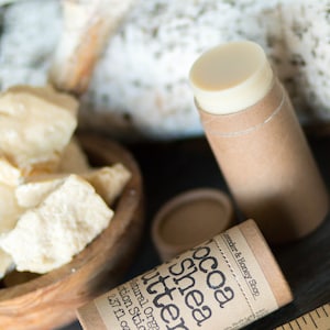 May include: A brown cardboard tube with a white label that reads "Cocoa Shea Butter Natural Organic Lotion Stick 2.37 fl oz" and "Lavender & Honey Shop". The tube is partially filled with a white lotion stick. There is a wooden ruler and a bowl of white shea butter in the background.