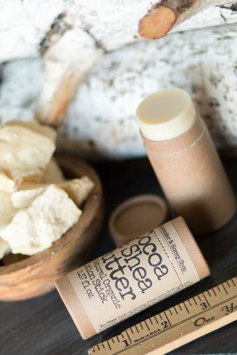 May include: A brown cardboard tube with a white label that reads "Cocoa Shea Butter Natural Organic Lotion Stick 2.37 fl oz" and "Lavender & Honey Shop". The tube is partially filled with a white lotion stick. There is a wooden ruler and a bowl of white shea butter in the background.