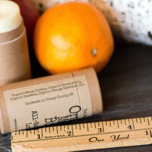 May include: A brown cardboard tube with a white lip balm inside. The tube has a label with the text "Organic Mango Butter, Organic Coconut Oil, Organic Beeswax, Organic Orange Essential Oil. Handmade in Orange County, CA. All-Natural Lip Balm. Orange Blossom"