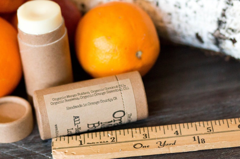 May include: A brown cardboard tube with a white lip balm inside. The tube has a label with the text "Organic Mango Butter, Organic Coconut Oil, Organic Beeswax, Organic Orange Essential Oil. Handmade in Orange County, CA. All-Natural Lip Balm. Orange Blossom"