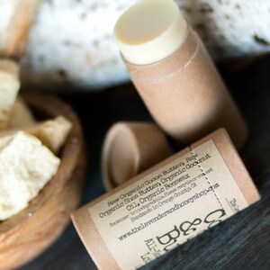 May include: A brown cardboard tube with a white label that reads "Raw Organic Cooon Buttery Rev Organic Shea Butter, Organic Coconut Oil, Organic Beeswax Container is biodegradable. Please recycle or compost. Handmade in Orange County, CA www.thelavenderandhoneyshop.com Lot 080". The tube is filled with a white, creamy substance.