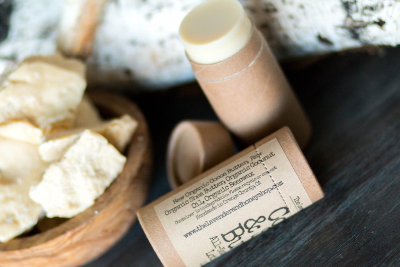 May include: A brown cardboard tube with a white label that reads "Raw Organic Cooon Buttery Rev Organic Shea Butter, Organic Coconut Oil, Organic Beeswax Container is biodegradable. Please recycle or compost. Handmade in Orange County, CA www.thelavenderandhoneyshop.com Lot 080". The tube is filled with a white, creamy substance.