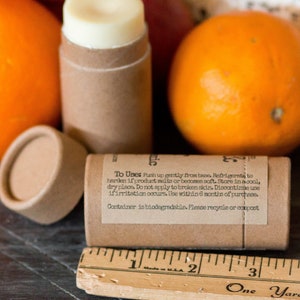 May include: A brown cardboard tube with a white lip balm inside. The tube has a label with the text "To Uses Push up gently from base. Refrigerate to harden if product melts or becomes soft. Store in a cool, dry place. Do not apply to broken skin. Discontinue use if irritation occurs. Use within 6 months of purchase. Container is biodegradable. Please recycle or compost." The tube is surrounded by oranges.