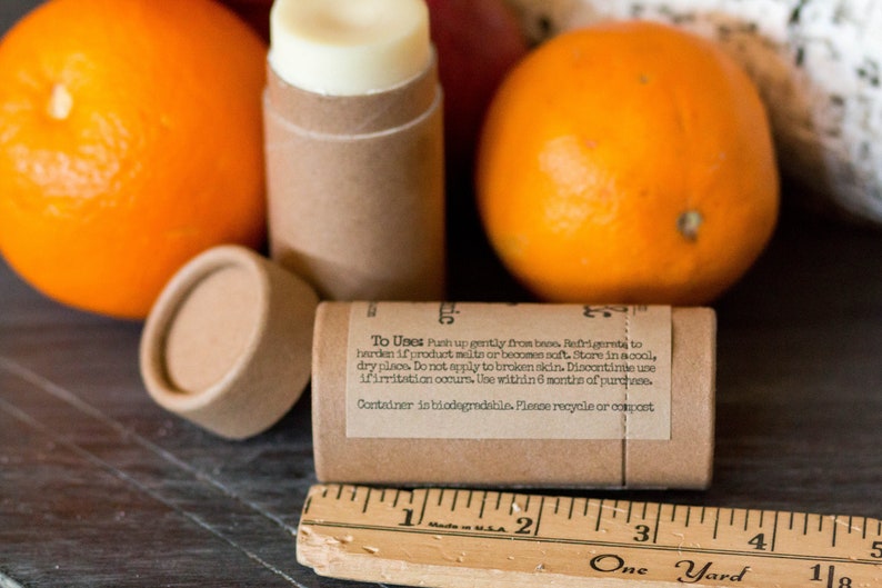 May include: A brown cardboard tube with a white lip balm inside. The tube has a label with the text "To Uses Push up gently from base. Refrigerate to harden if product melts or becomes soft. Store in a cool, dry place. Do not apply to broken skin. Discontinue use if irritation occurs. Use within 6 months of purchase. Container is biodegradable. Please recycle or compost." The tube is surrounded by oranges.