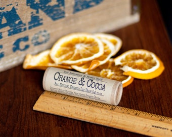 May include: A lip balm tube with the text "Orange & Cocoa" and dried orange slices are displayed on a wooden surface. A wooden ruler is next to the lip balm. The lip balm is labeled "All Natural Organic Lip Balm 0.5 fl oz."
