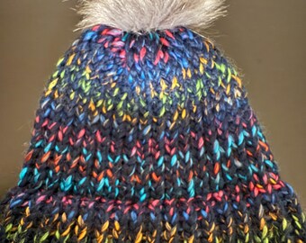 May include: A colorful knit beanie with a fluffy pom pom on top. The beanie is made with a variety of colors, including black, blue, green, yellow, and orange.
