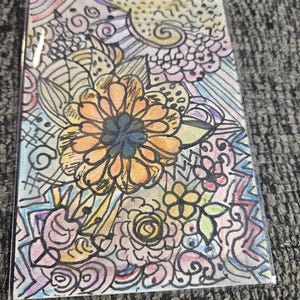 May include: A colorful floral art print featuring a large orange flower with a dark blue center, surrounded by smaller flowers and abstract patterns. The artwork is encased in a clear plastic sleeve, showcasing a vibrant mix of colors and intricate details.