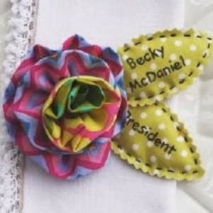 May include: A yellow fabric flower with a pink, blue, and green chevron pattern. The flower is attached to a yellow fabric leaf with the text "Becky McDaniel President" embroidered in black.