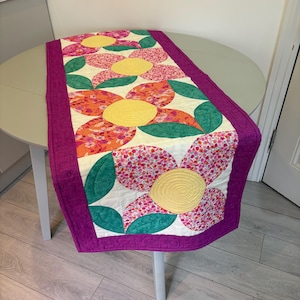May include: A rectangular quilted table runner featuring a cream background with a repeating floral pattern of large yellow circular centers, pink and orange floral-print petals, and green leaves, all bordered by a solid purple fabric trim.