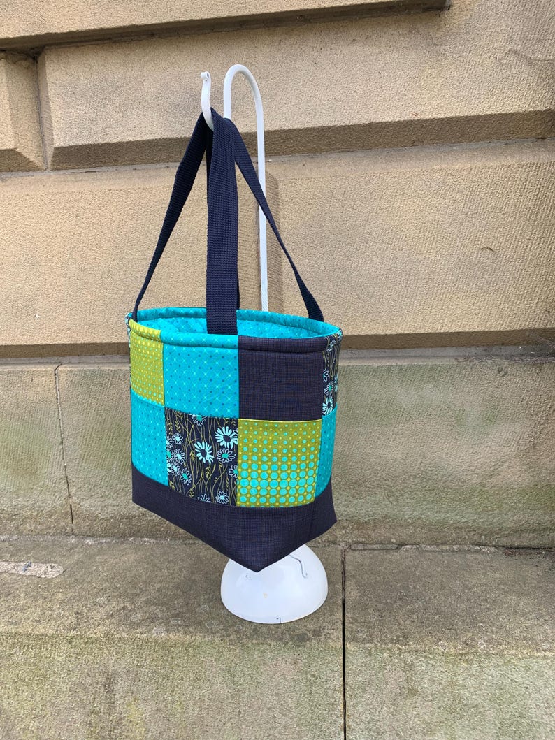 K&ouml;nnte beinhalten: Eine Patchwork-Tragetasche mit marineblauem Griff und Besatz. Die Tasche hat Quadrate aus t&uuml;rkisem, gr&uuml;nem und floralem Stoff. Die Tasche h&auml;ngt an einem wei&szlig;en Haken vor einer Steinmauer.