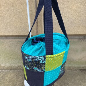 K&ouml;nnte beinhalten: Eine Patchwork-Tragetasche mit marineblauem Griff und t&uuml;rkisblauem Innenraum. Die Au&szlig;enseite weist eine Mischung aus gemusterten Stoffen in T&uuml;rkis, Gr&uuml;n und Marineblau auf. Die Tasche h&auml;ngt an einem wei&szlig;en Haken.