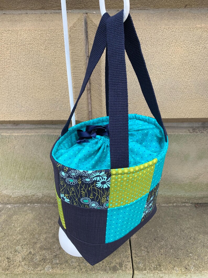 K&ouml;nnte beinhalten: Eine Patchwork-Tragetasche mit marineblauem Griff und t&uuml;rkisblauem Innenraum. Die Au&szlig;enseite weist eine Mischung aus gemusterten Stoffen in T&uuml;rkis, Gr&uuml;n und Marineblau auf. Die Tasche h&auml;ngt an einem wei&szlig;en Haken.