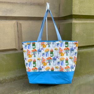 May include: A floral tote bag with a white background and a pattern of colorful flowers in vases and watering cans. The bag has a bright blue bottom and light blue handles, perfect for carrying groceries or other items.