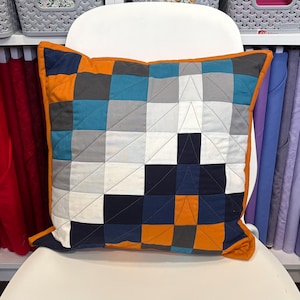 May include: Square patchwork pillow with a geometric design in shades of blue, gray, white, and orange. The pillow has an orange border and is placed on a white chair. The pillow is a decorative home accent.