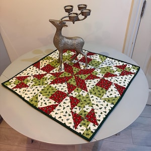 May include: A decorative Christmas table runner with a green border, featuring a starburst pattern in red, green, and white fabric. A silver deer-shaped candle holder with four candle cups sits atop the runner, creating a festive holiday display.