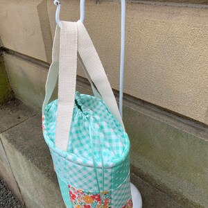 K&ouml;nnte beinhalten: Eine Patchwork-Tragetasche mit Kordelzugverschluss, die eine Mischung aus floralen, Vichy- und einfarbigen t&uuml;rkisfarbenen Stoffen aufweist. Die Tasche hat einen wei&szlig;en Riemen und ist an einem wei&szlig;en Metallst&auml;nder aufgeh&auml;ngt. Das Design der Tasche ist eine Mischung aus Mustern und Farben.