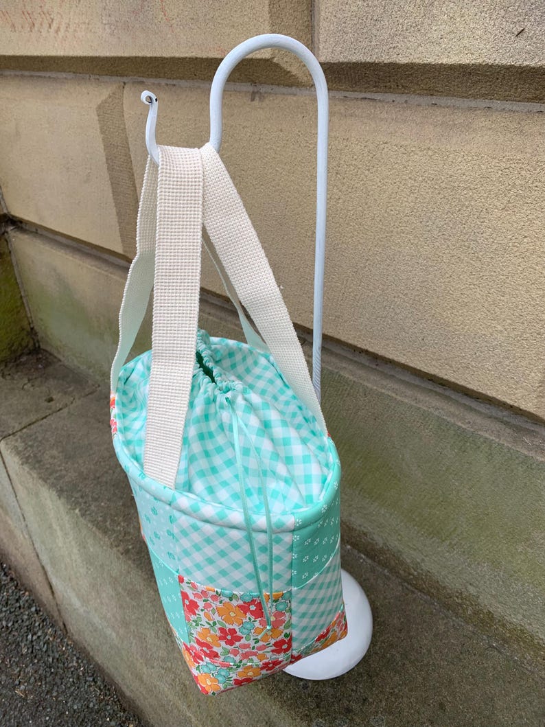 K&ouml;nnte beinhalten: Eine Patchwork-Tragetasche mit Kordelzugverschluss, die eine Mischung aus floralen, Vichy- und einfarbigen t&uuml;rkisfarbenen Stoffen aufweist. Die Tasche hat einen wei&szlig;en Riemen und ist an einem wei&szlig;en Metallst&auml;nder aufgeh&auml;ngt. Das Design der Tasche ist eine Mischung aus Mustern und Farben.