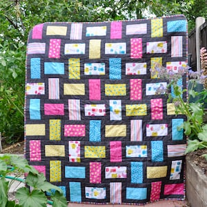 Könnte beinhalten: Ein farbenfroher Patchwork-Deckenbezug mit schwarzem Hintergrund und einer Vielzahl von rechteckigen Blöcken in rosa, gelben, blauen und weißen Stoffen. Die Decke ist mit einer Vielzahl von Mustern gefertigt, darunter Punkte, Streifen und Blumenmuster.