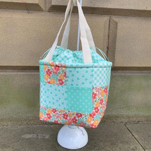 K&ouml;nnte beinhalten: Eine Patchwork-Tragetasche mit Kordelzug und wei&szlig;en Griffen. Die Tasche hat eine Mischung aus Blumen-, Tupfen- und Vichy-Mustern in T&uuml;rkis-, Orange- und Wei&szlig;t&ouml;nen. Die Tasche h&auml;ngt an einem wei&szlig;en Haken.