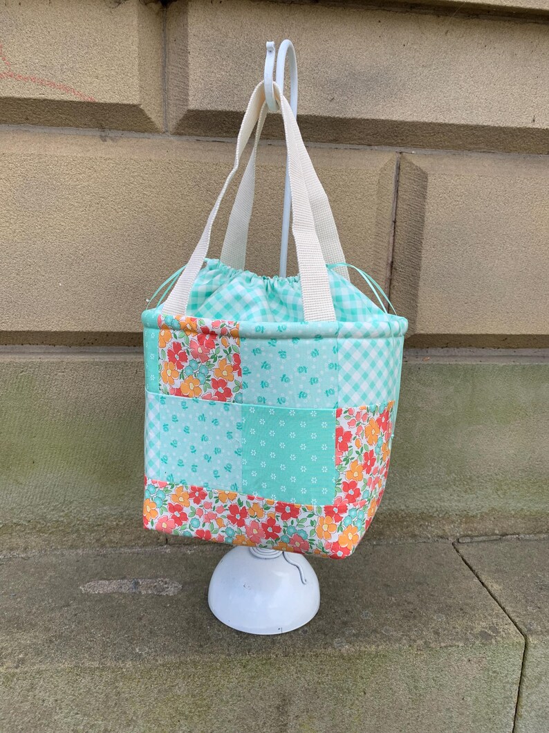 K&ouml;nnte beinhalten: Eine Patchwork-Tragetasche mit Kordelzug und wei&szlig;en Griffen. Die Tasche hat eine Mischung aus Blumen-, Tupfen- und Vichy-Mustern in T&uuml;rkis-, Orange- und Wei&szlig;t&ouml;nen. Die Tasche h&auml;ngt an einem wei&szlig;en Haken.