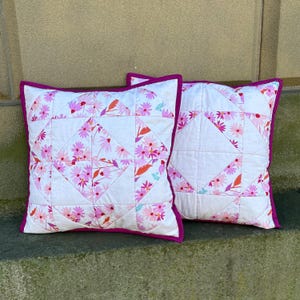 May include: Two square throw pillows with white fabric featuring a floral pattern in pink, orange, and green. The pillows have a quilted design with a magenta border. The pillows are placed on a stone surface.