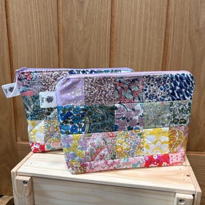May include: Two patchwork zipper pouches with floral patterns in various colors. The pouches are rectangular with a light purple zipper and a small tag attached. They are sitting on a wooden box.