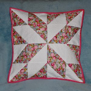 Op de afbeelding: Een wit kussen met een roze en witte stippenrand. Het kussen is gemaakt van stof met een bloemenpatroon en is gestikt in een stervorm.