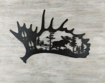 May include: Black metal silhouette of a moose walking through a forest, cut out of a moose antler shape.