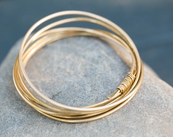 May include: A set of gold-colored bangles, resting on a gray stone. The bangles are thin and circular, with a wrapped wire detail at one point. The jewelry is simple and elegant, suitable for everyday wear.