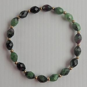 Puede incluir: Un brazalete de piedras preciosas verdes y rojas con cuentas doradas. El brazalete está hecho de cuentas ovaladas facetadas.