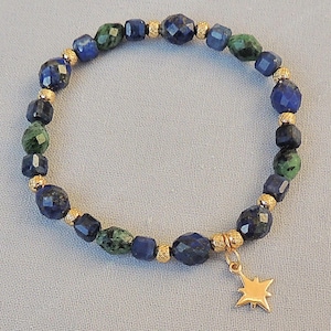 May include: A beaded bracelet featuring faceted blue and green gemstones interspersed with gold-colored beads. A gold star charm hangs from the bracelet. The bracelet is displayed on a light gray surface.