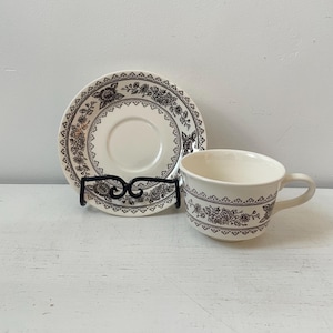 May include: Cream-colored teacup and saucer set with a floral pattern in dark brown. The cup has a handle and the saucer is round. The set is displayed on a white surface.
