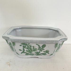 May include: White ceramic planter with an octagonal shape and a green floral design. The planter has a decorative border and is suitable for indoor or outdoor use. The planter is empty.