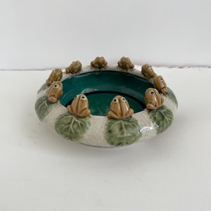 May include: A ceramic bowl with a dark green interior and a white exterior decorated with brown frog figurines. The bowl's rim is adorned with small, detailed frogs, and the exterior features green leaf designs.