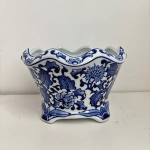 May include: A white ceramic planter featuring a blue floral pattern with intricate botanical details. The vessel has a scalloped, wavy rim and stands on four small, curved feet, showcasing a classic blue and white color scheme throughout.
