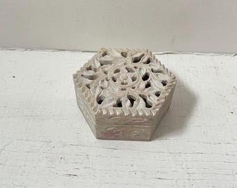 May include: A light beige, hexagonal box with intricate floral carvings on the lid. The box has a detailed, openwork design, and the sides are smooth. The box is made of a stone-like material and sits on a white, distressed surface.