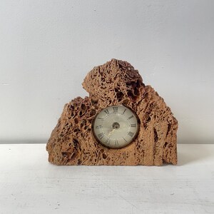 May include: A brown, textured clock with a round face featuring Roman numerals. The clock is set within a uniquely shaped, porous, rock-like structure. The clock hands indicate the time. The overall design is rustic and natural.