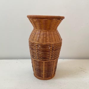 May include: A tall, woven wicker vase in shades of brown. The vase has a flared top and a cylindrical body, with a textured, basket-weave pattern. The vase is approximately 18 inches tall.