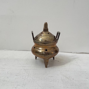 May include: A small, antique brass incense burner with a lid and three legs. The burner has a rounded body with etched floral designs and small holes for releasing incense smoke. The lid has a small handle and two side handles.