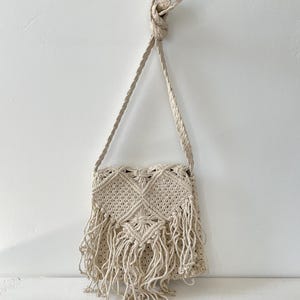 May include: Cream-colored macrame shoulder bag with a woven design and fringe detail. The bag features a diamond pattern in the center and a braided strap. The bag is hanging against a white background.