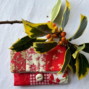 May include: A patchwork wallet with a button closure, featuring red and cream floral and gingham patterns. The wallet is accented with a sprig of holly with yellow-edged green leaves and orange berries.