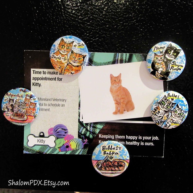 May include: Five colorful round magnets with painted images of cats. The magnets feature different cat breeds and names, including Zayde, Tate, Mame, Shayna, Punim, and Bubbe. The magnets are on a black background with a white card that says "Time to make an appointment for Kitty." and "Keeping them happy is your job. Keeping them healthy is ours."