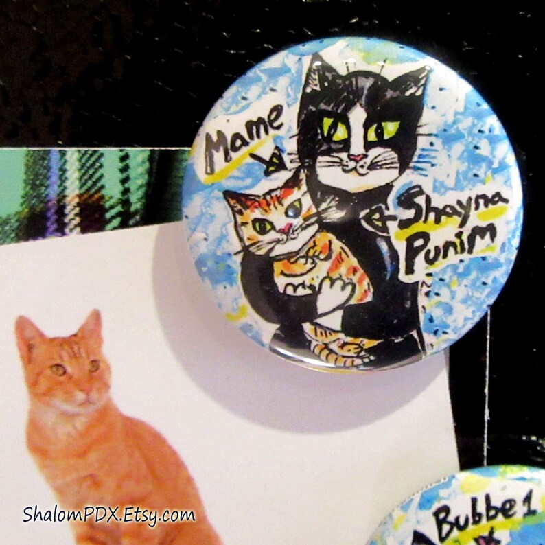 May include: A blue and white button with a black and white illustration of two cats. The larger cat is holding a smaller cat. The text "Mame & Shayna Punim" is written on the button.