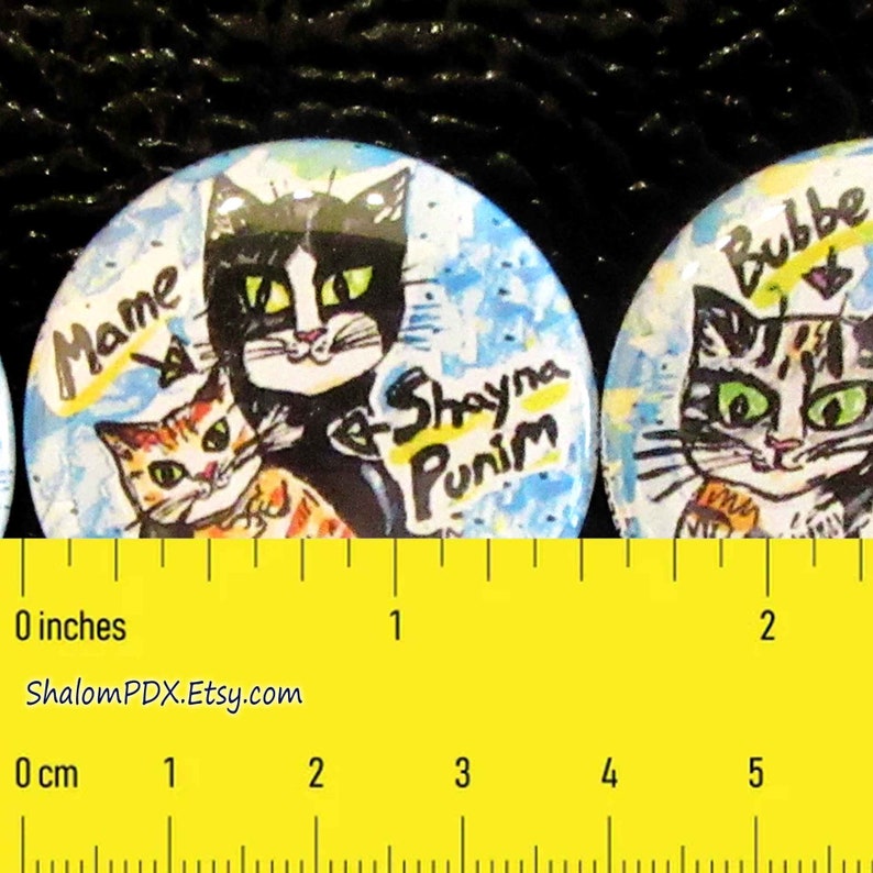 May include: Two round buttons with colorful cat illustrations and text. The first button features a black and white cat with green eyes and the text "Mame & Shayna Punim". The second button features a tabby cat with green eyes and the text "Bubbe".